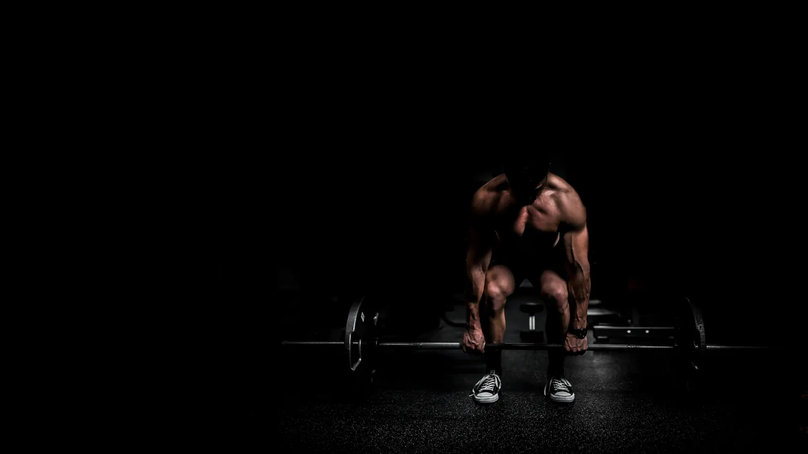 Persona levantando una barra olímpica desde el piso bajo un foco de luz, en un gimnasio a oscuras